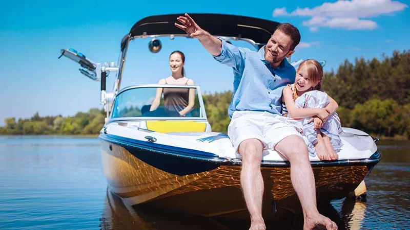 a dad sits on the front of a ship with his daughter while mom drives behind at the wheel