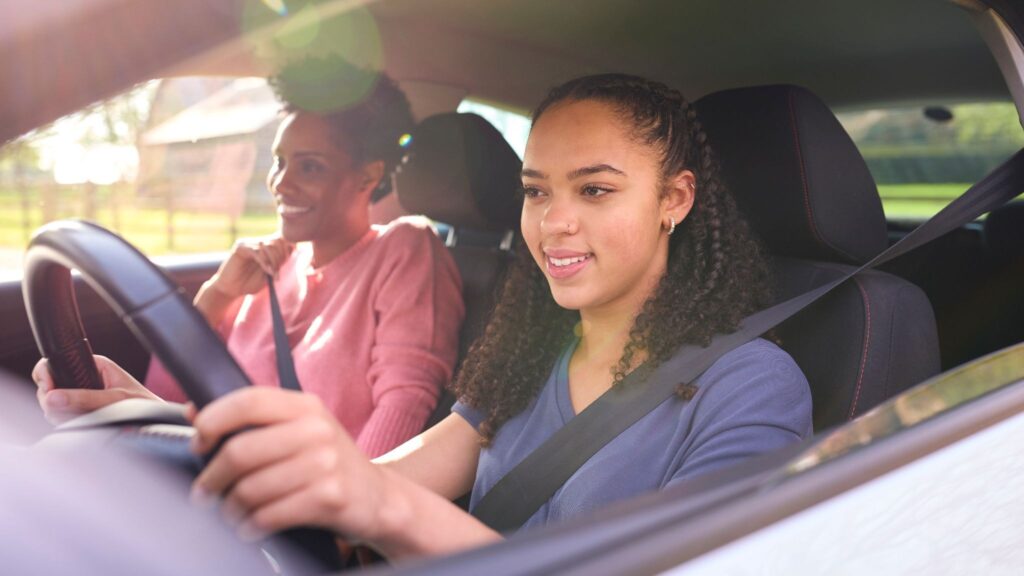 Parent standing with a teenage driver next to a family car, representing the higher insurance costs and increased risk associated with adding a new teen driver to an auto insurance policy.